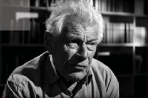 Close-up black and white photo of an elderly man with white hair, in a room with bookshelves in the background.