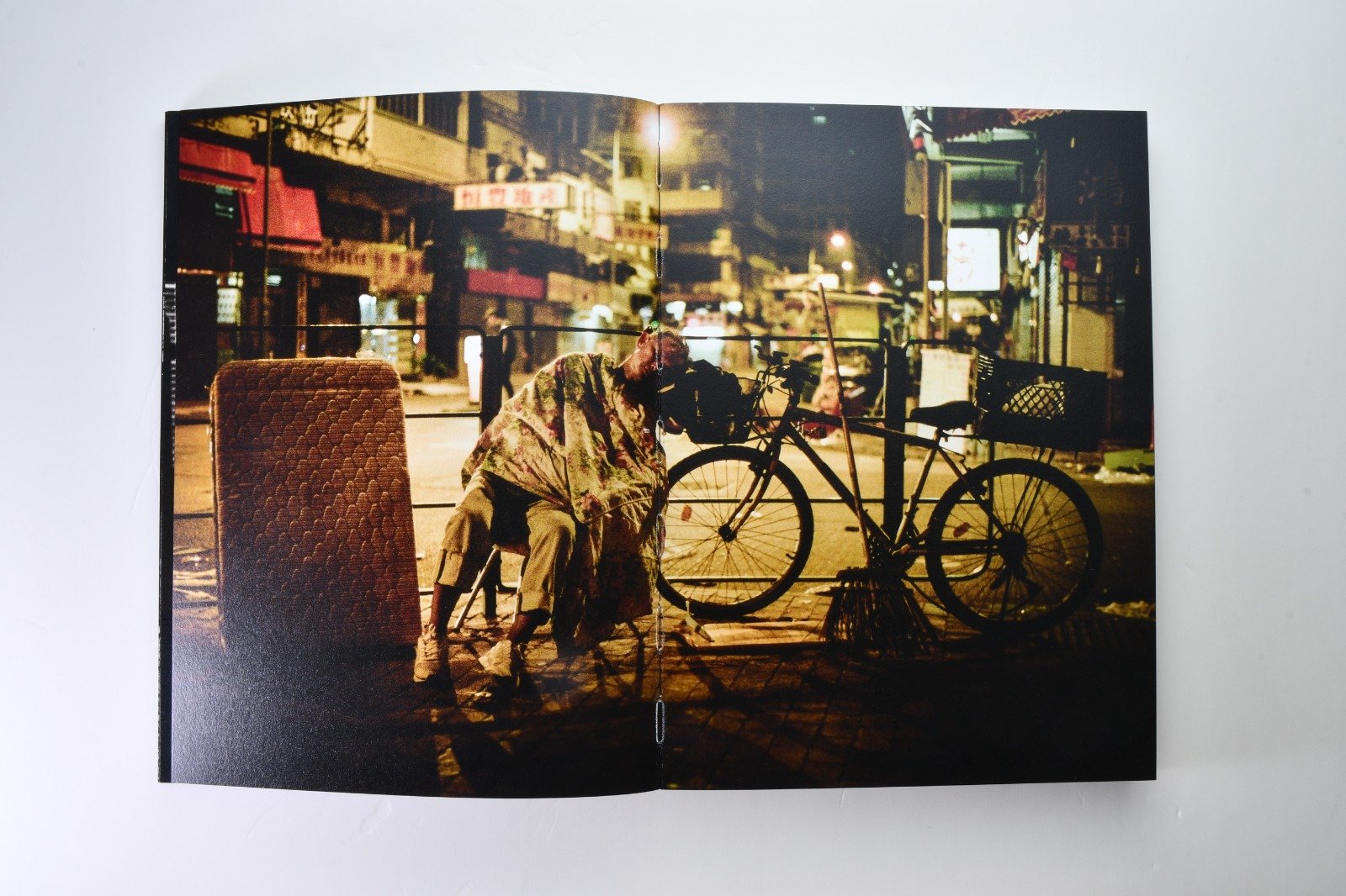 A homeless person sleeps on the sidewalk at night next to a bicycle, with city buildings and streetlights in the background.