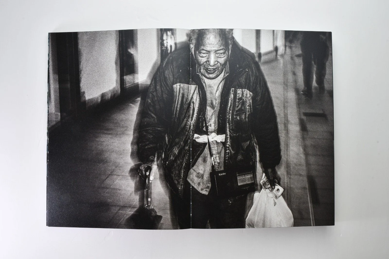 Black and white photo of an elderly man with a cane, carrying a shopping bag and a camera, walking indoors.