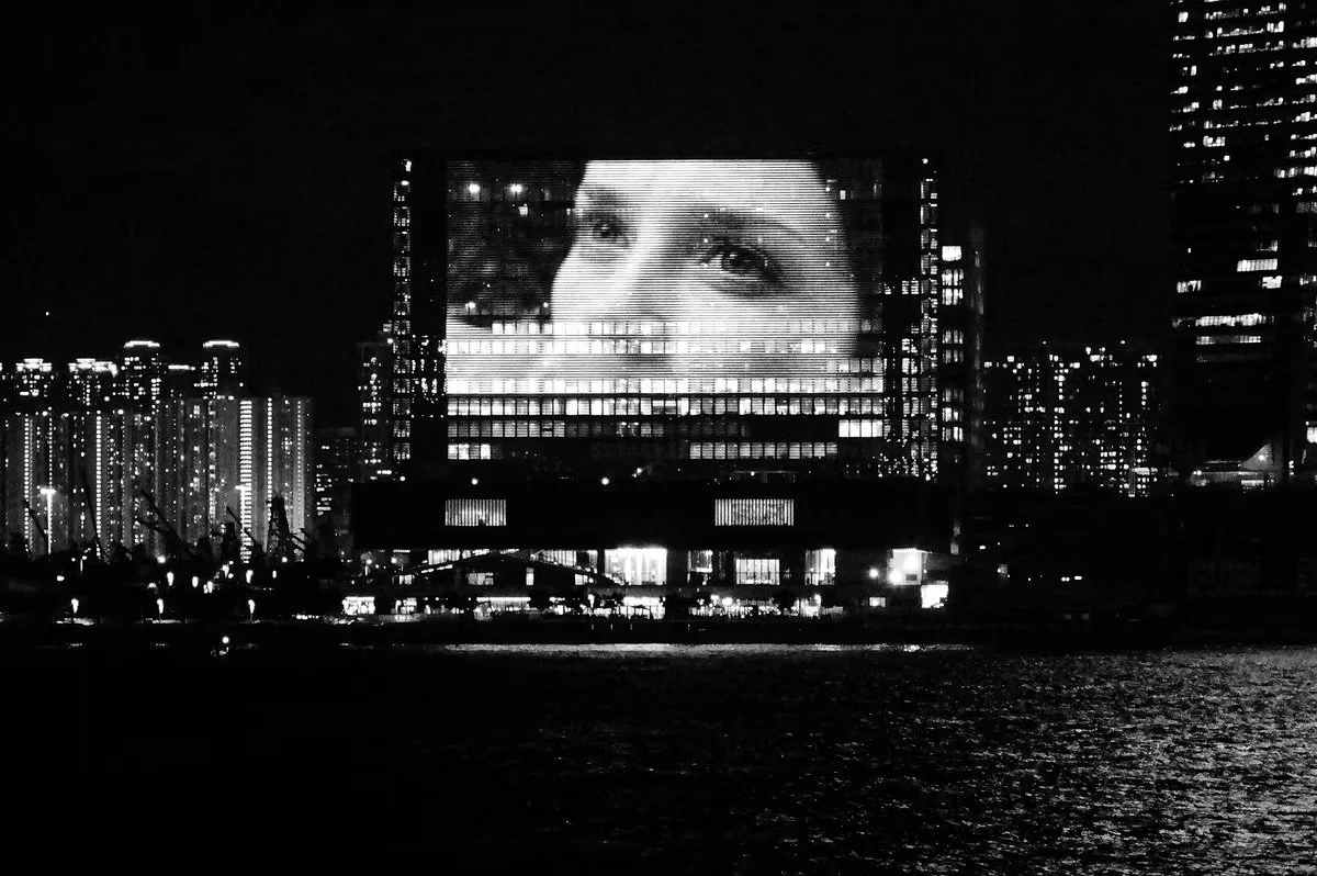 Nighttime cityscape with a large digital billboard displaying a woman's face, surrounded by illuminated buildings.