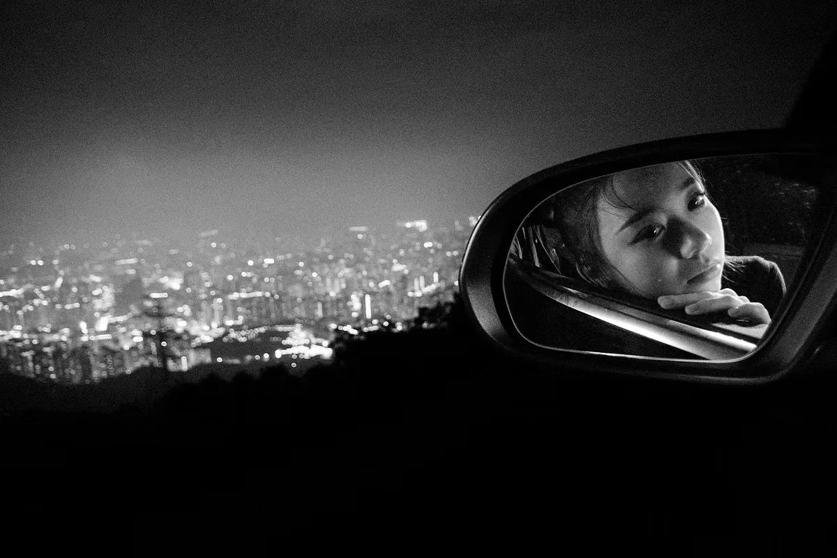 Black and white photo of a young woman looking into a car's side mirror at night, cityscape with lights in the background.