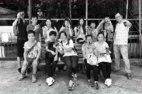 Group of kids and teenagers outdoors, some sitting on a bench, others standing, with trees and a fence in the background.