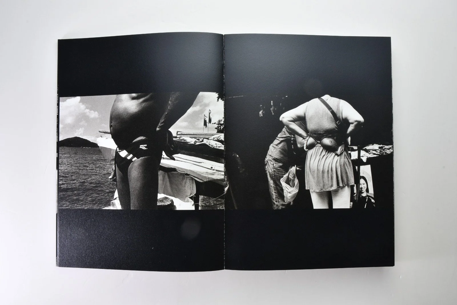 Black and white photograph of people bending over a table outdoors, near a body of water, with a cloudy sky in the background.