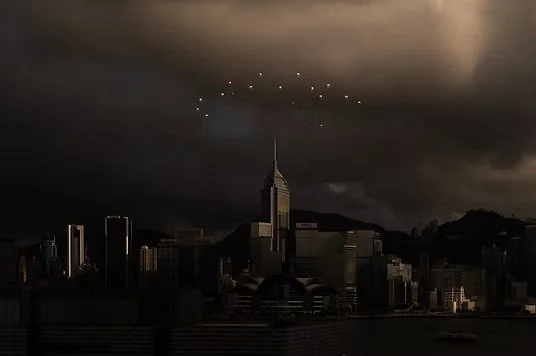 Dark city skyline with the Empire State Building, dark clouds, and a formation of drones or lights flying in a curve overhead.