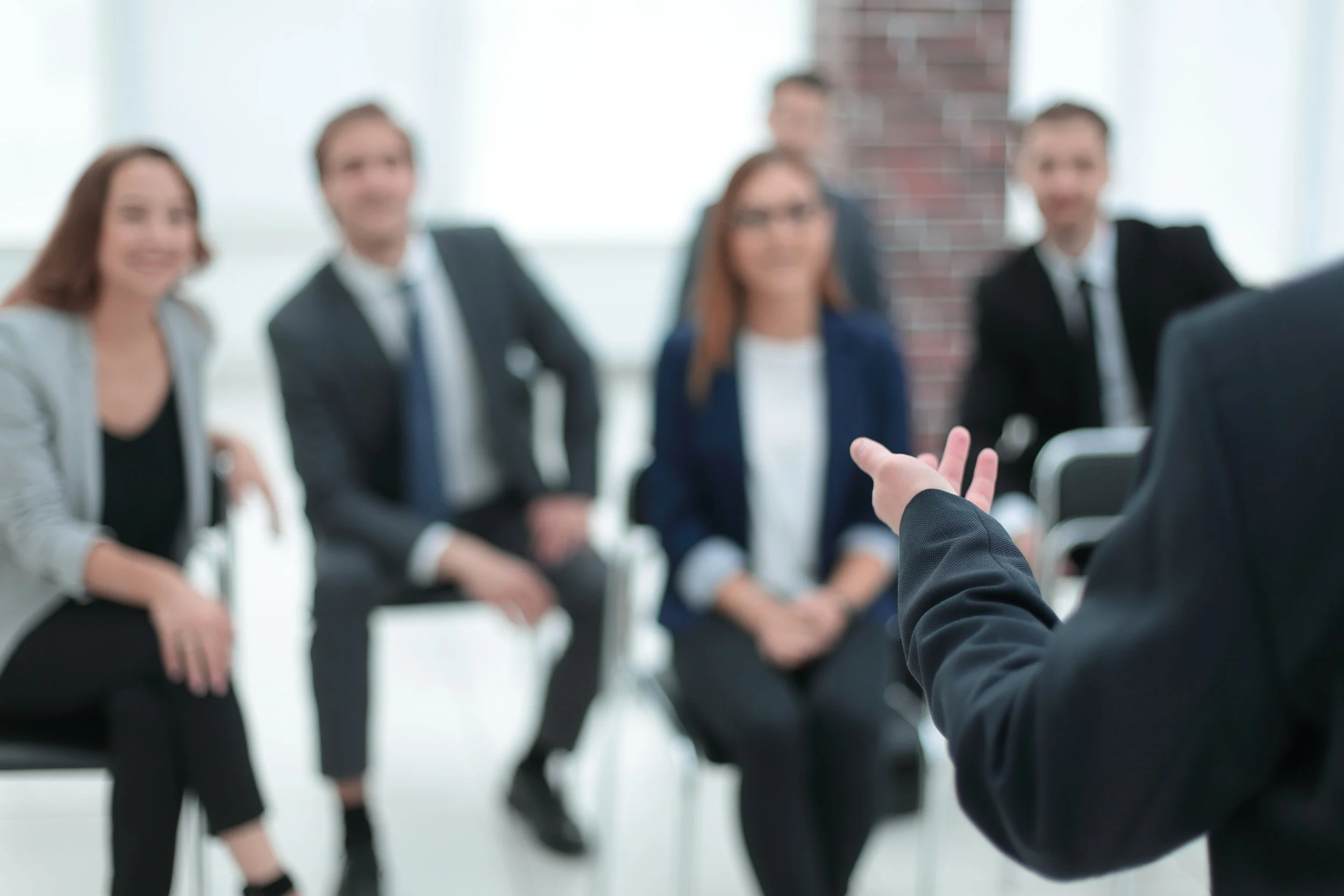 A group of five people in business attire sitting in chairs, blurred background, listening to an executive coach.