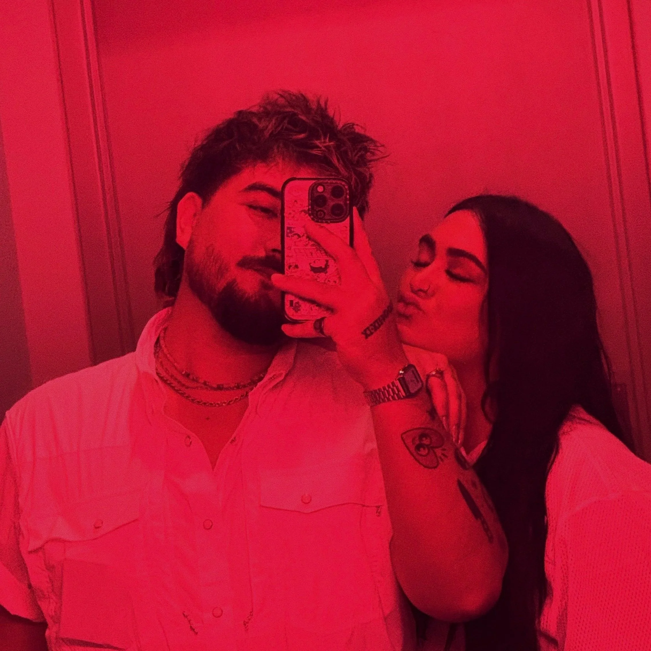 A person with short, spiked hair taking a selfie in front of a mirror with a smartphone. The person is wearing a dark top and accessories. Another person with long hair, also wearing glasses, is partially visible next to them, leaning in for the photo. The background shows a plain wall with decorative molding.