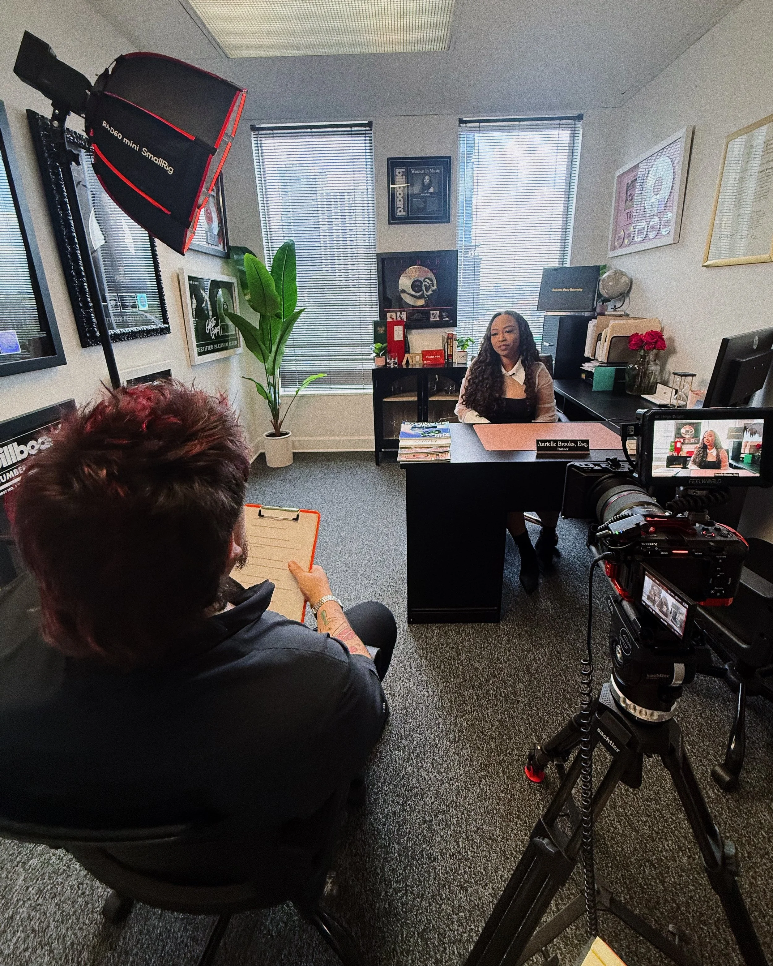 An office with a woman sitting at a desk, speaking to someone off-camera. The office has multiple framed pictures on the walls, a large potted plant, and shelves with books and decorations. A professional camera on a tripod is set up in the foreground.