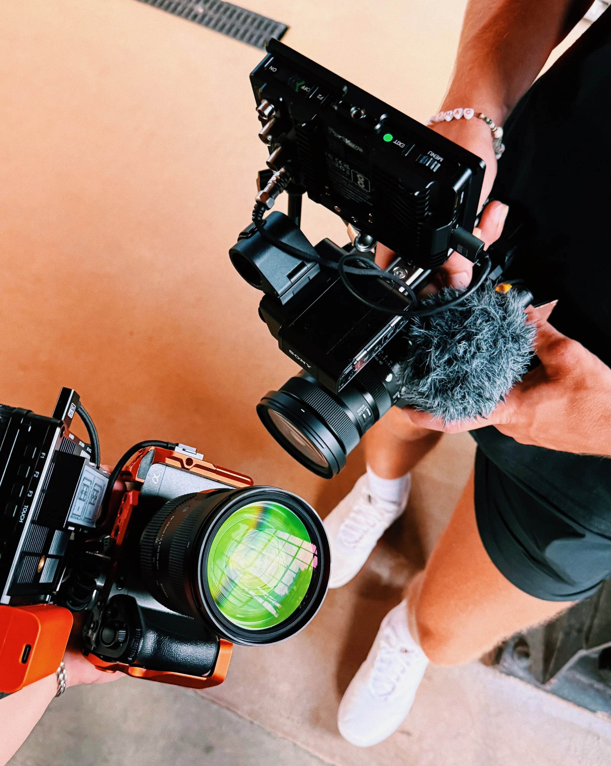 Camera mounted on a stabilizer held by a person wearing white sneakers, black shorts, and a bead bracelet, with other camera equipment visible on a beige surface.