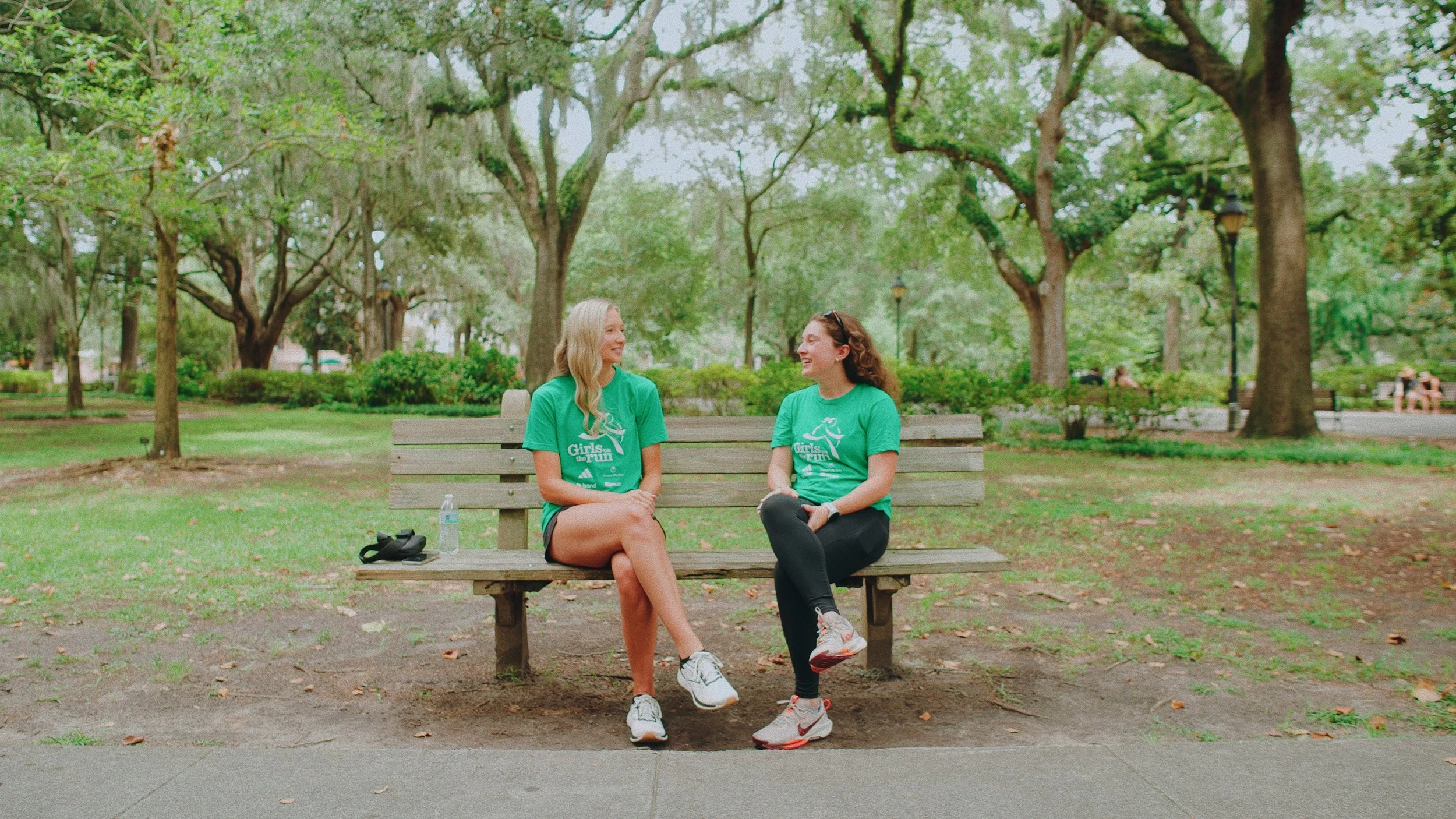 Two women sitting on a park bench, smiling and talking, surrounded by green trees and grass