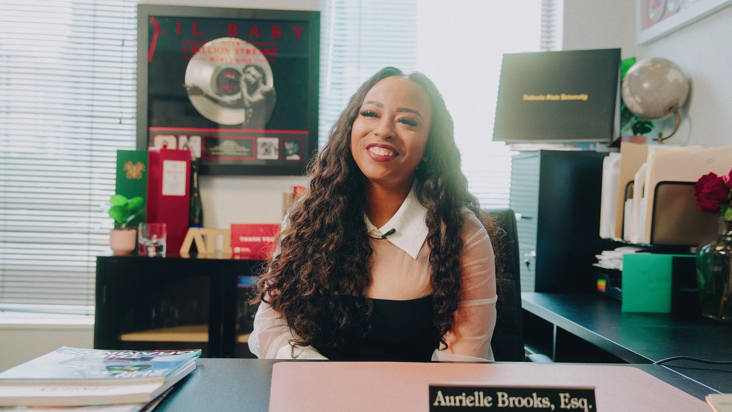 A woman with long, curly hair sitting at a desk in an office, smiling, with a nameplate that reads 'Aurielle Brooks, Esq.' in front of her. Behind her are office supplies, a globe, and a framed poster of Lil Baby.
