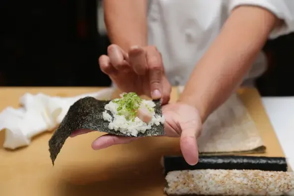 Pessoa preparando um sushi, colocando recheio de peixe e cebolinha em uma folha de alga nori.