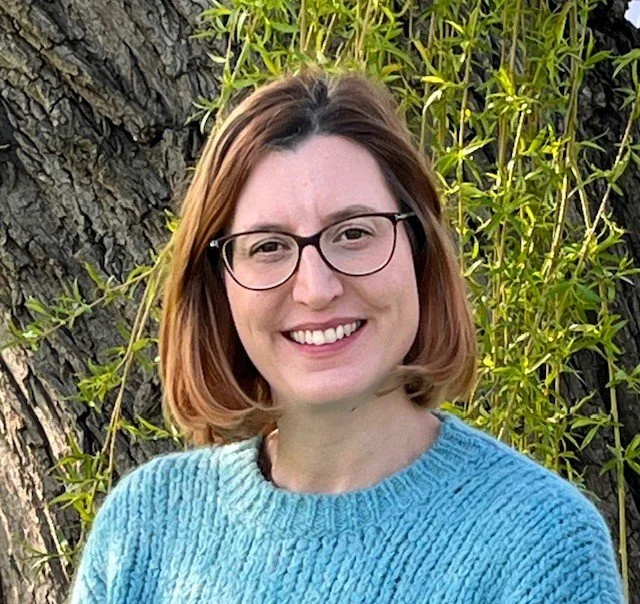A woman with shoulder-length brown hair, glasses, and a blue sweater, smiling outdoors in front of a willow tree with green leaves.