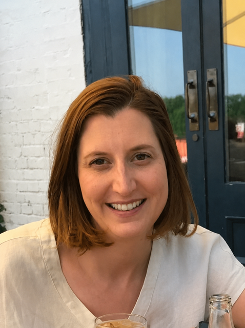 A woman with short red hair smiling at the camera, wearing a light-colored shirt, sitting outside near a blue door with glass panels, in front of a white brick wall.