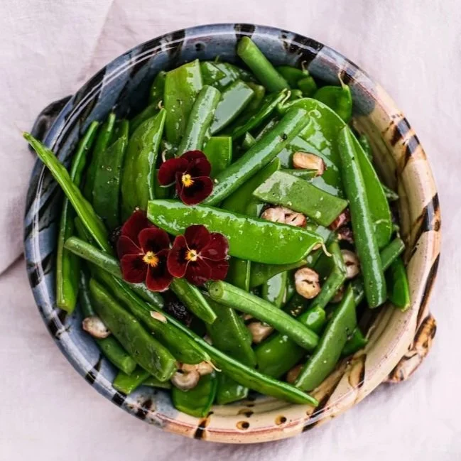 Fresh green sugar snap peas in a container, garnished with edible purple flowers and small garlic cloves.