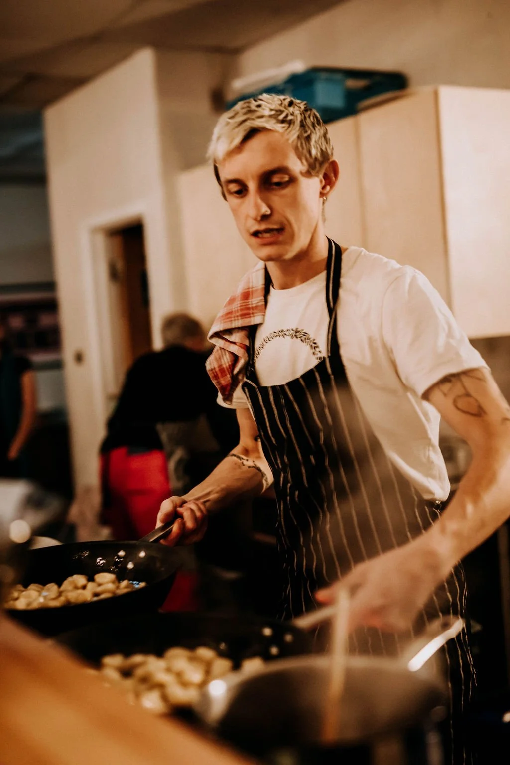 Young man with blond hair cooking food in a restaurant kitchen, wearing a black and white striped apron and a white t-shirt, with a scarf over his shoulder.