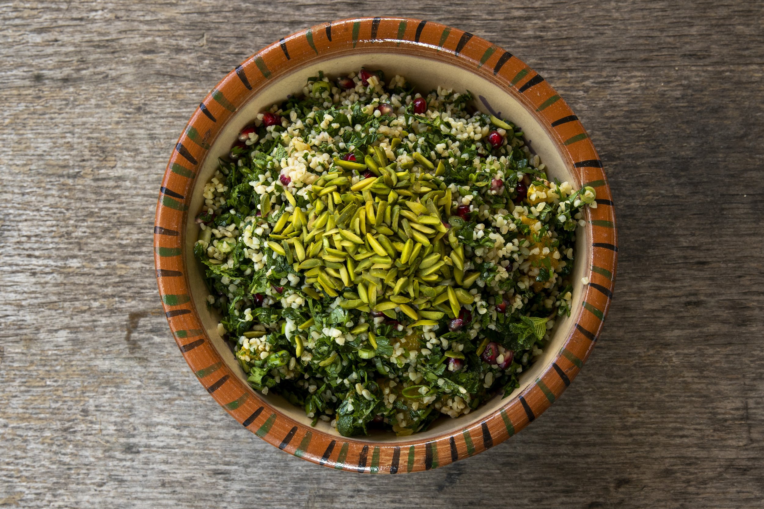A bowl of tabbouleh salad with bulgur, parsley, mint, pomegranate seeds, and sunflower seeds on a rustic wooden surface.
