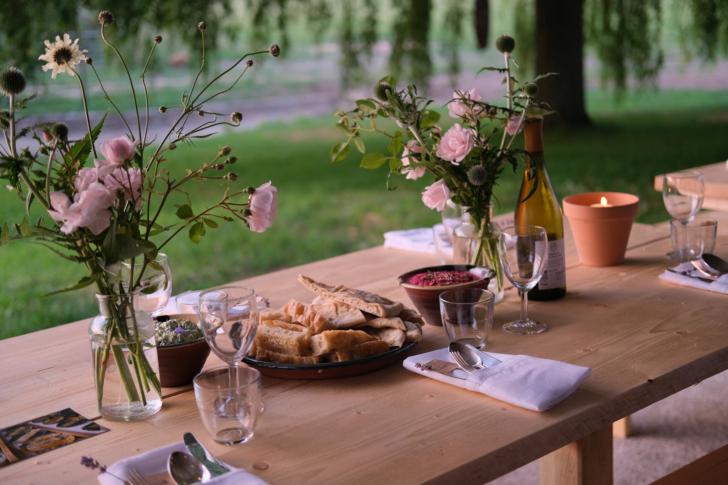 A wooden table set outdoors with flower arrangements, bread, dips, glasses, and a lit candle, against a background of green trees.