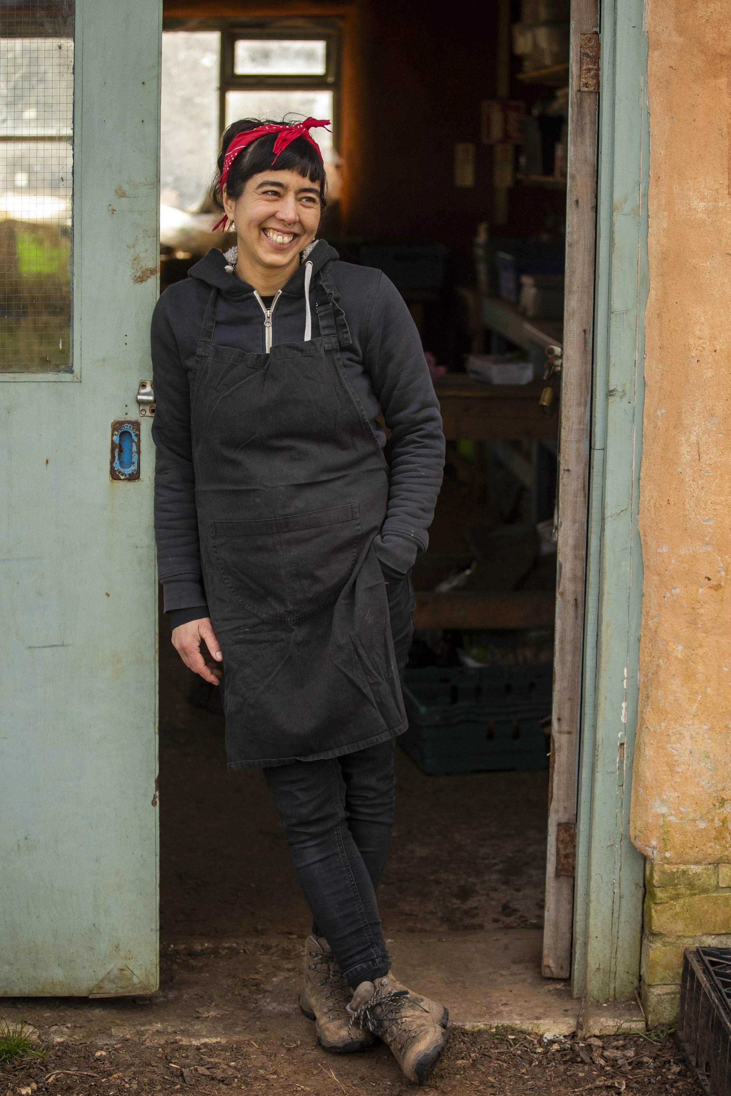 A woman standing in a doorway, wearing a red headband, black hoodie, apron, black pants, and hiking boots, smiling and looking off to the side inside a rustic building.