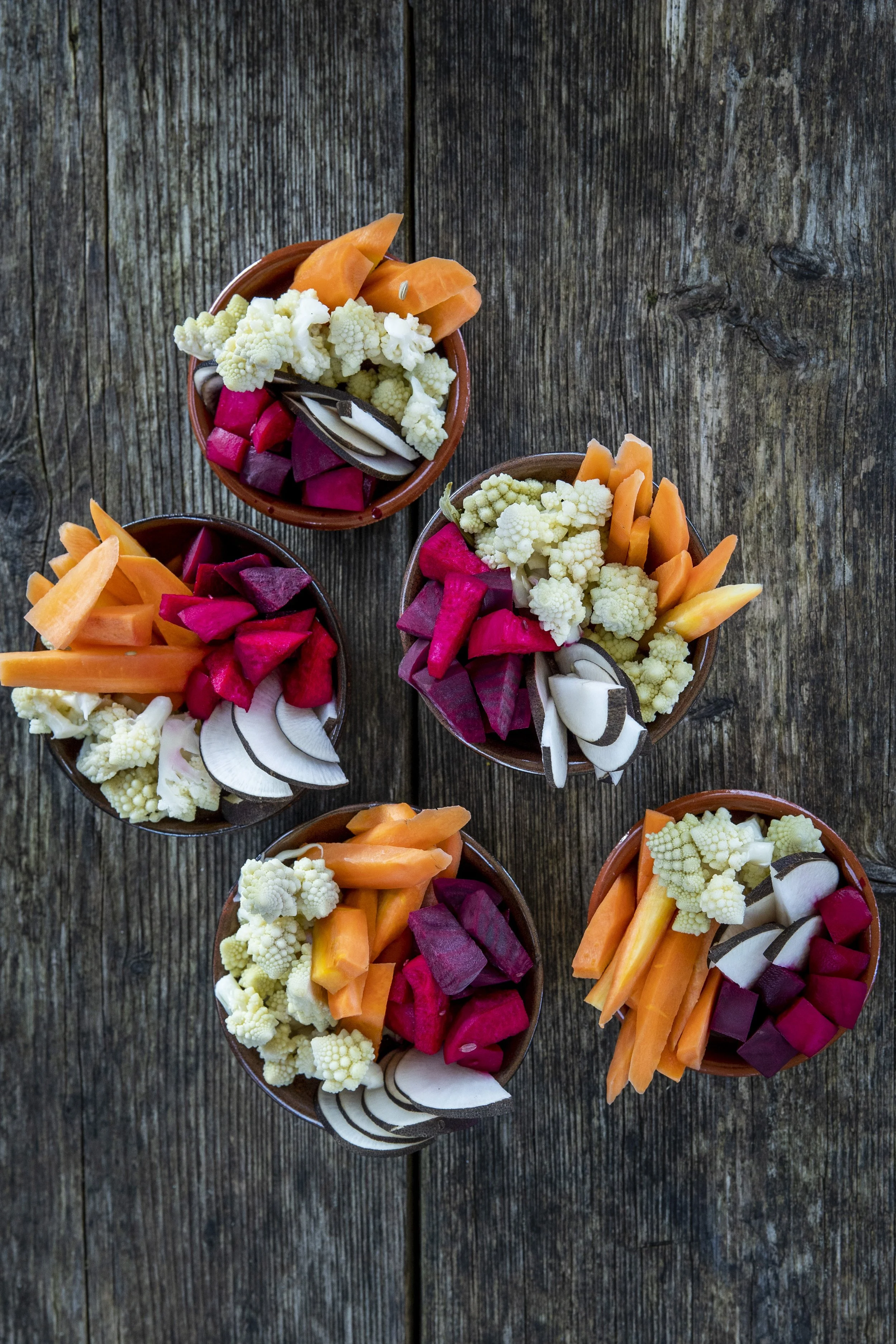 Multiple small bowls filled with an assortment of sliced vegetables including cauliflower, carrots, radishes, and purple beets, arranged on a rustic wooden surface.