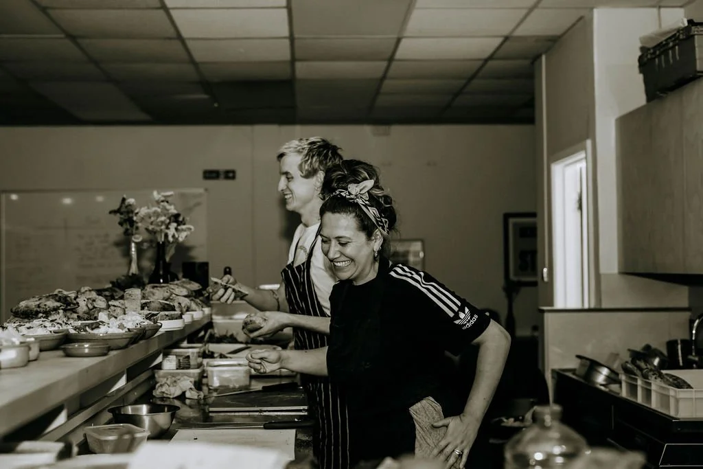 Two women in a kitchen laughing and preparing food. One woman is wearing a black Adidas shirt with white stripes, and the other is wearing a striped apron. There are various bowls and plated food on the countertop.