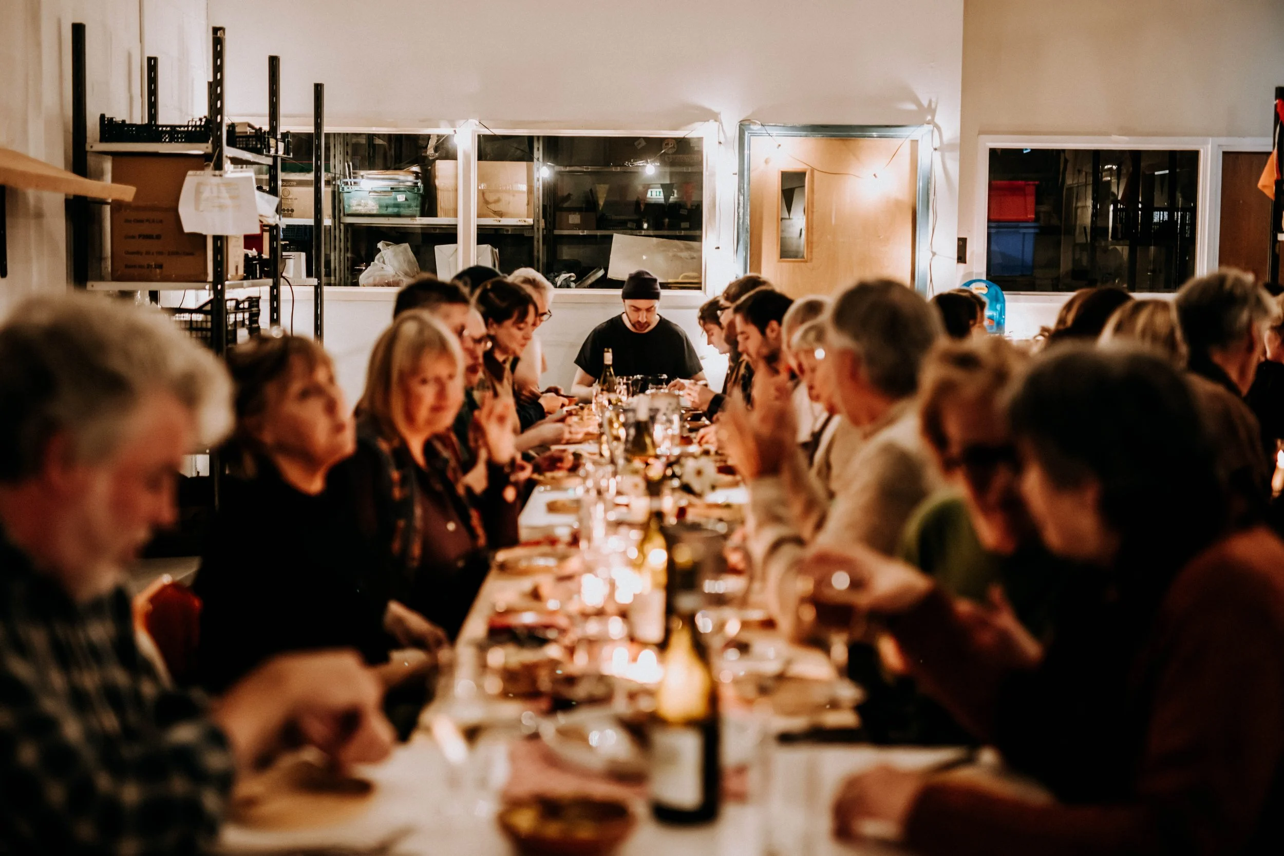 People sitting at a long table dining together in a cozy indoor setting with warm lighting and large windows.