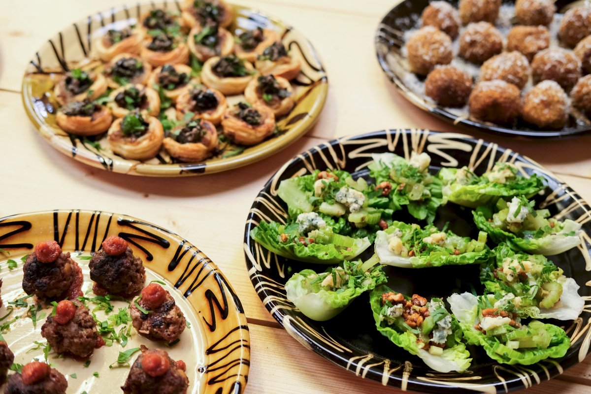 A variety of appetizer dishes on decorative plates, including stuffed endive leaves with blue cheese and walnuts, meatballs with tomato sauce, a plate of fried balls coated in powdered sugar, and a plate of pinwheel appetizers