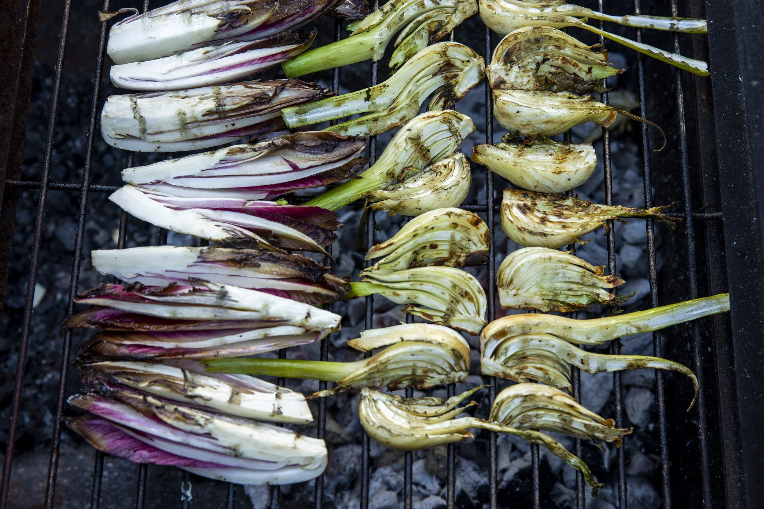 Vegetables being grilled on a barbecue grill. The vegetables include endives and radicchio, some of which are charred.
