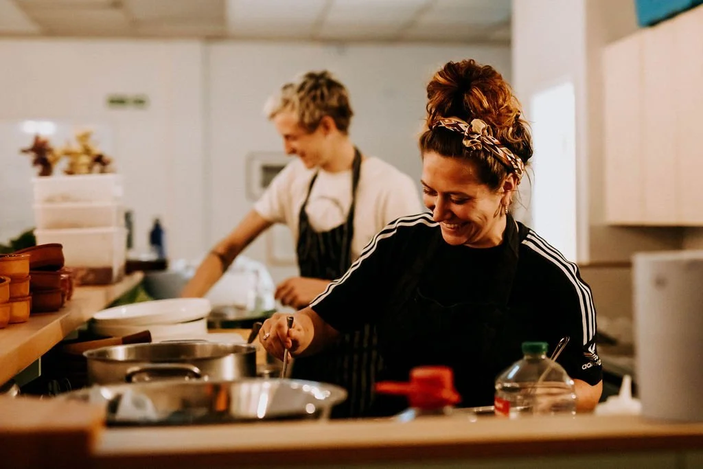 Smiling woman with a headband cooking in a kitchen, man in the background preparing food.