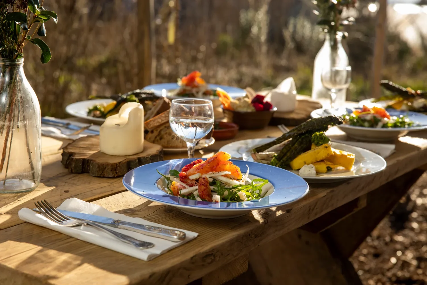 An outdoor wooden table set with various dishes, including salad, grilled asparagus, and bread, with a lit candle and water glasses, in natural sunlight.