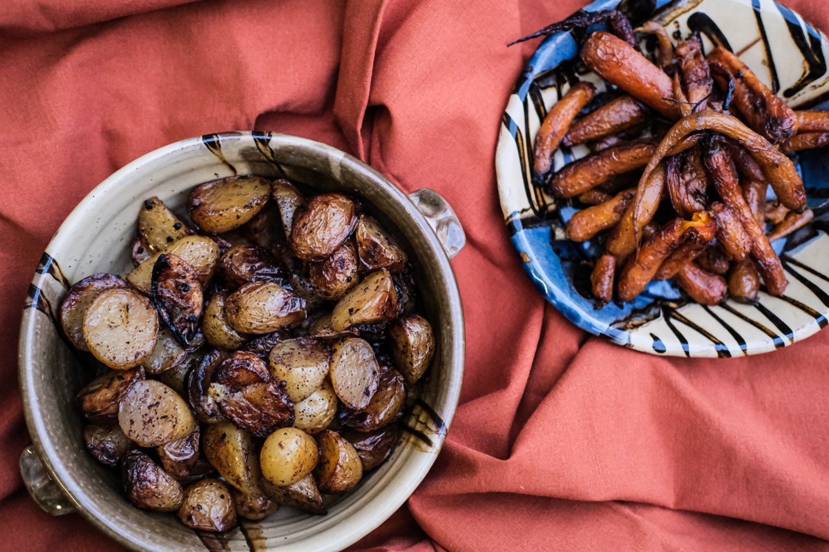 Two bowls of cooked baby potatoes, one seasoned and roasted, the other glazed with caramelized carrots, on a reddish cloth.