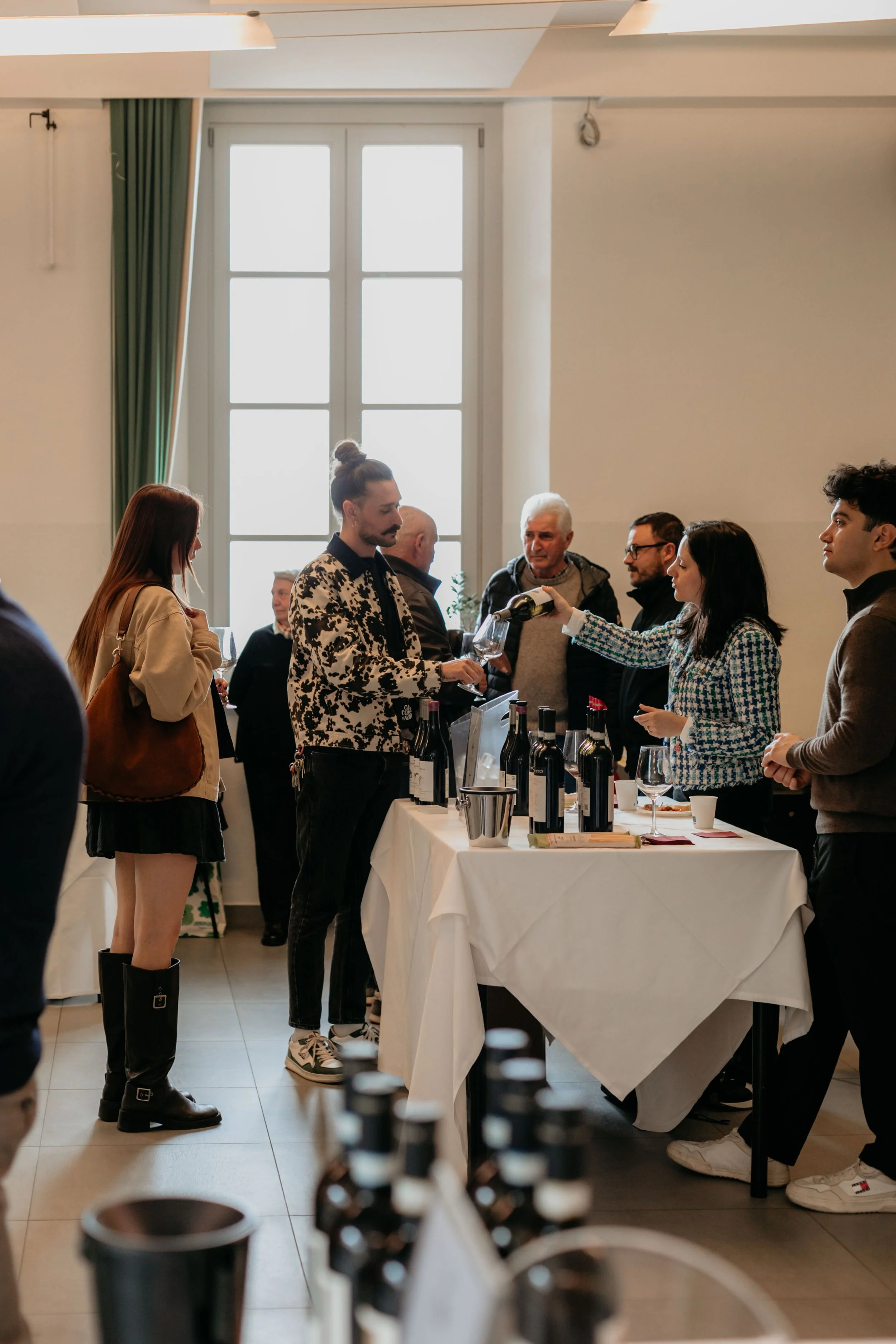 Gruppo di persone che degustano vino durante un evento in una stanza luminosa con grandi finestroni.
