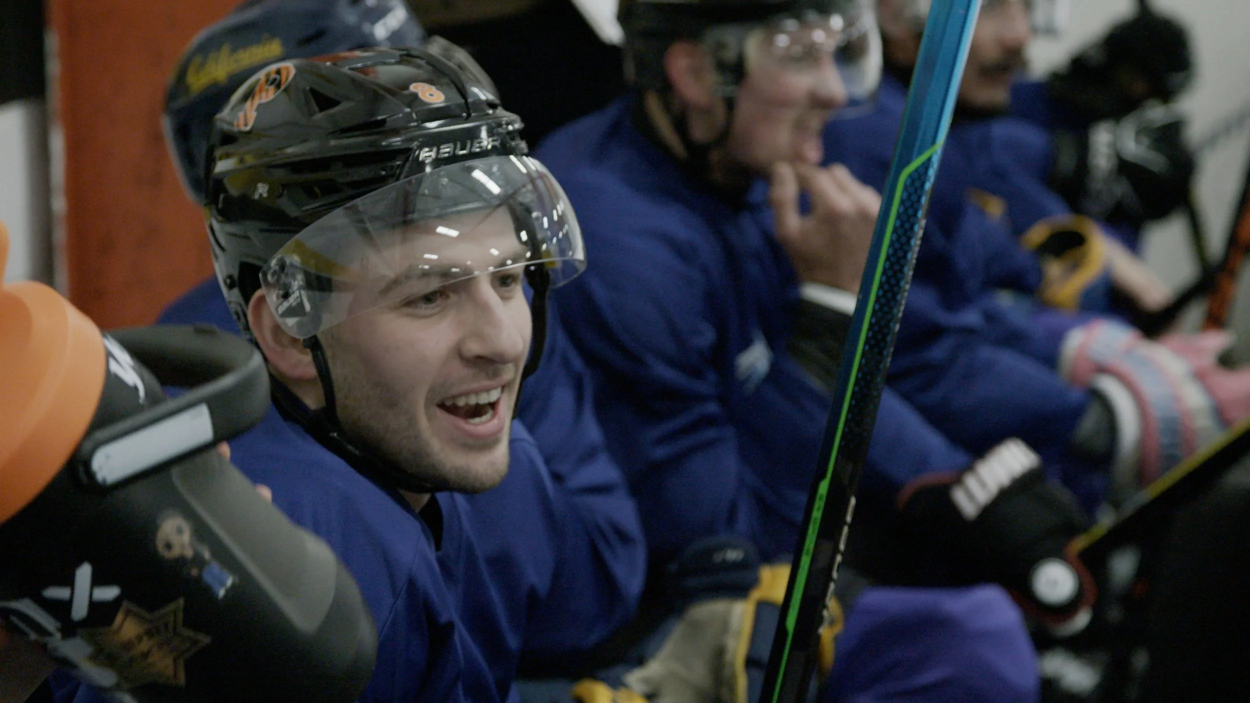 Hockey players sitting on the bench, smiling, wearing blue uniforms and helmets, with hockey sticks, in an indoor ice rink.