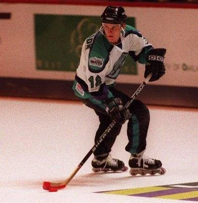 A hockey player in a white and teal uniform with number 12, wearing a helmet and gloves, skating on an ice rink with a hockey stick in hand.