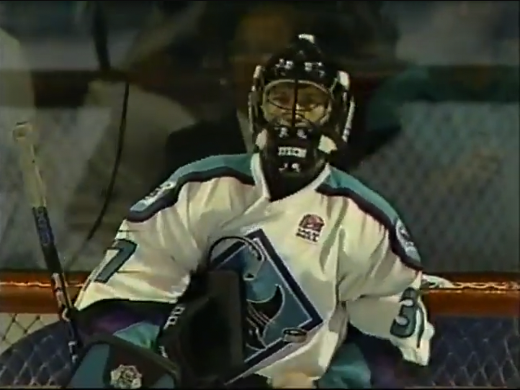 Hockey goalie in full gear, wearing a white jersey with teal and black accents, and a face mask, sitting in front of the hockey net.