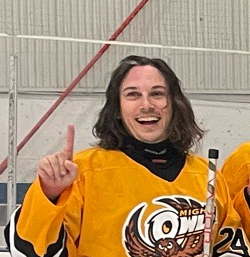 Young woman in a yellow ice hockey jersey smiling and pointing upward inside an ice rink