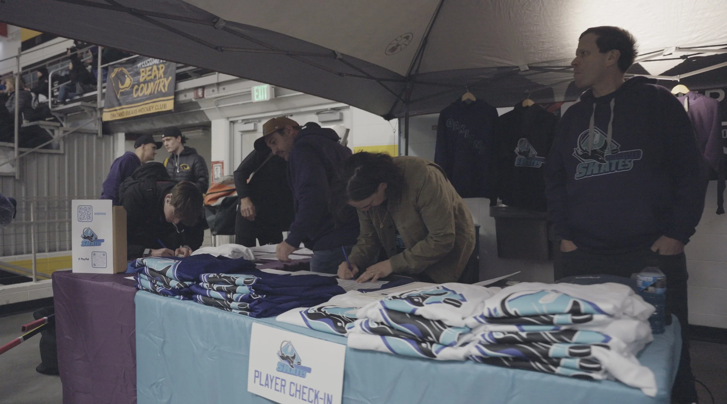 People at a table signing documents and collecting hockey jerseys at a Player Check-In booth under a tent, with Ottawa Bears signage and hockey apparel in the background.