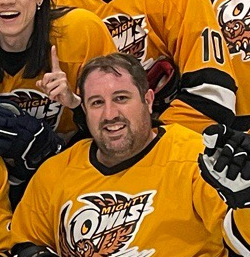 A man in a yellow hockey jersey smiling and giving a thumbs-up, surrounded by teammates in similar jerseys.