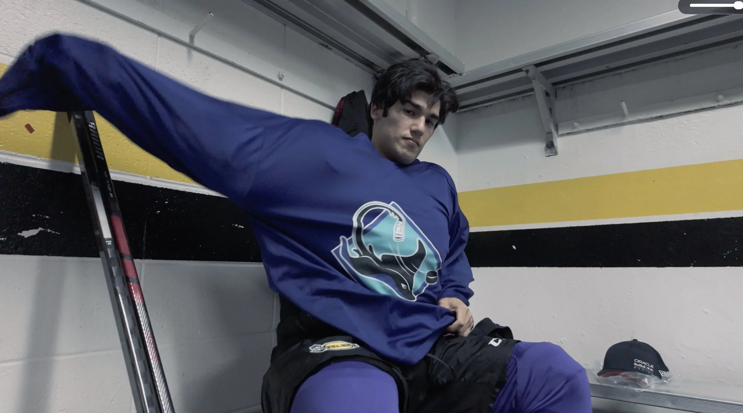 A young man sitting on a bench in a hockey locker room, wearing a blue hockey jersey with a logo on the front, black hockey pants, and blue hockey socks. He has dark hair and is looking at the camera, with a hockey stick leaning against the wall next to him and a cap on the shelf beside him.