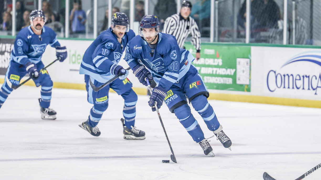 Hockey players in blue uniforms skating on ice rink during a game, with referee in the background.