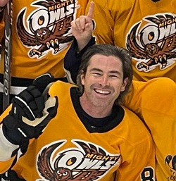 Hockey player smiling and making a peace sign, wearing a yellow jersey, with teammates in the background.