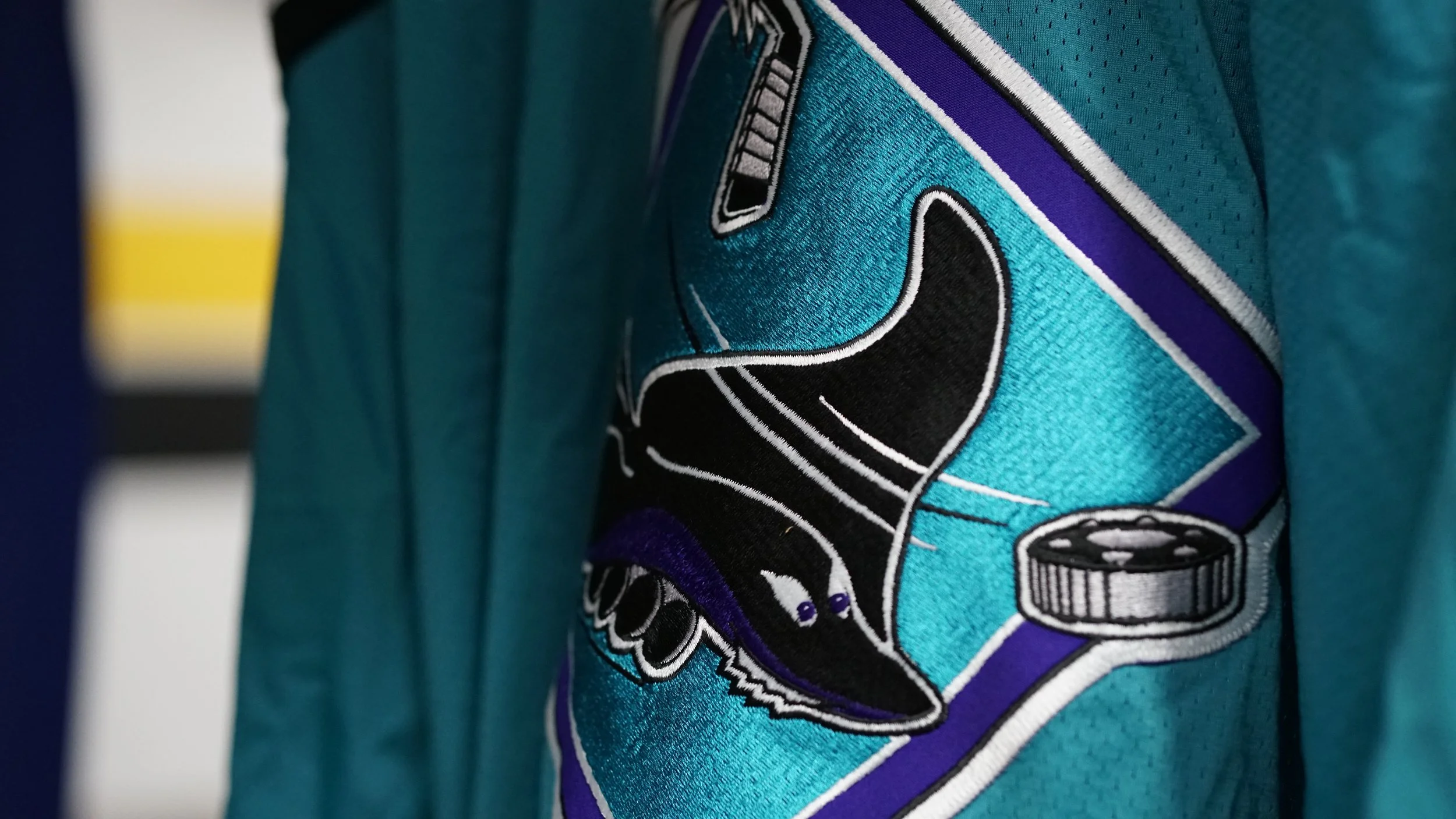 Close-up of a teal hockey jersey with an embroidered San Jose Sharks logo, featuring a black shark with purple and white accents, and a hockey puck.