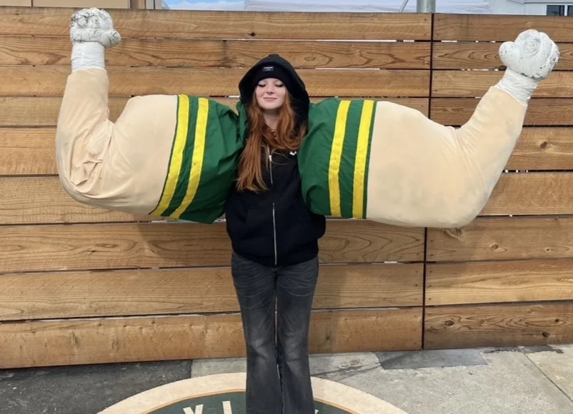 Person standing in front of a wooden fence, posing with oversized green and beige exaggerated muscular arms attached to a statue or costume.