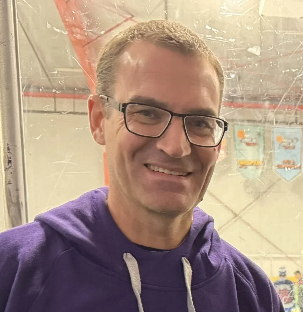 Smiling man with glasses and purple hoodie in an indoor ice hockey rink.
