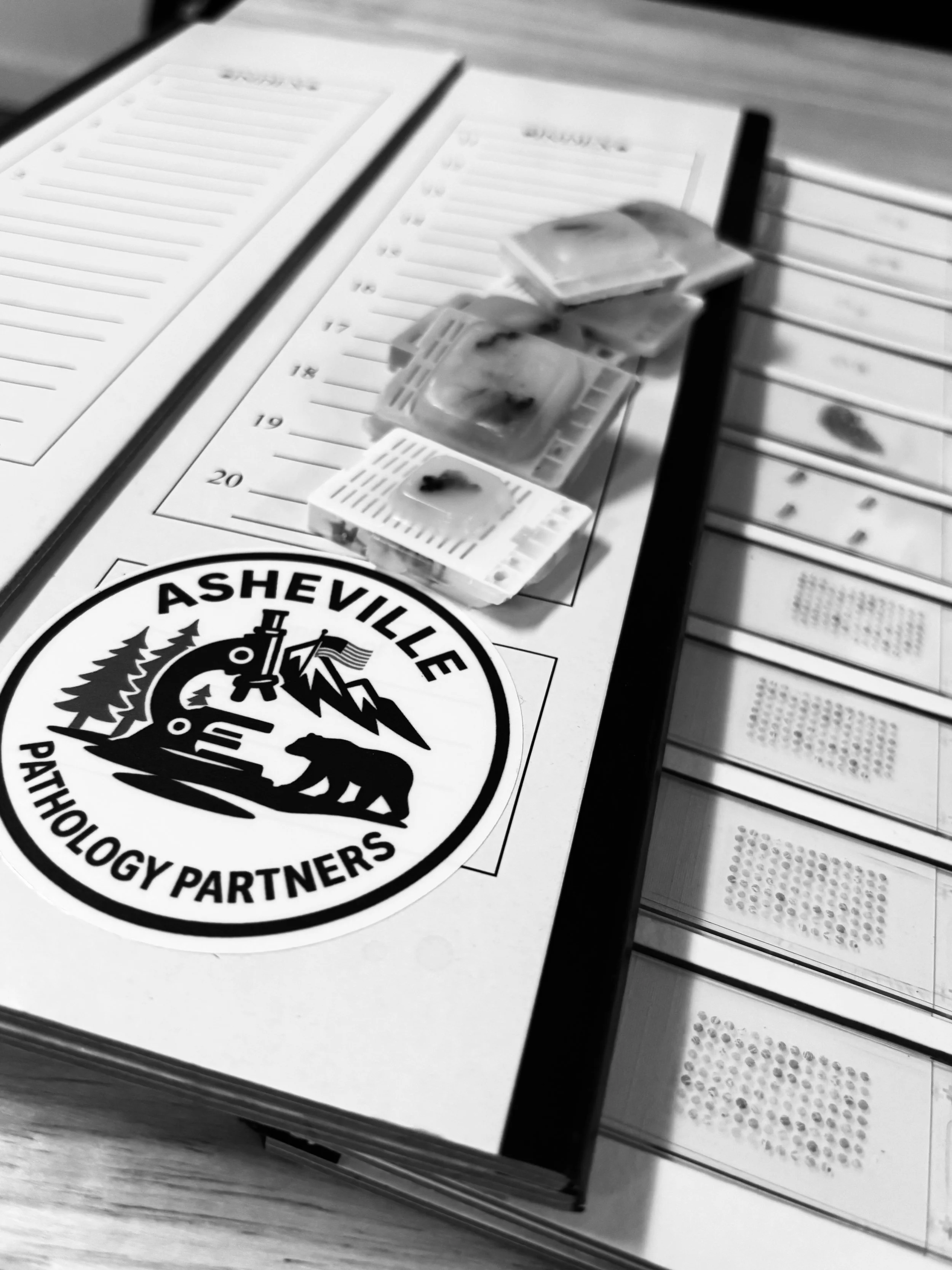 B&W photo of slide folders with microscope slides that have a 102 core tissue microarray as well as some routine slides. Tissue blocks are also on top of the slide folders with the Asheville Pathology Partners logo present.
