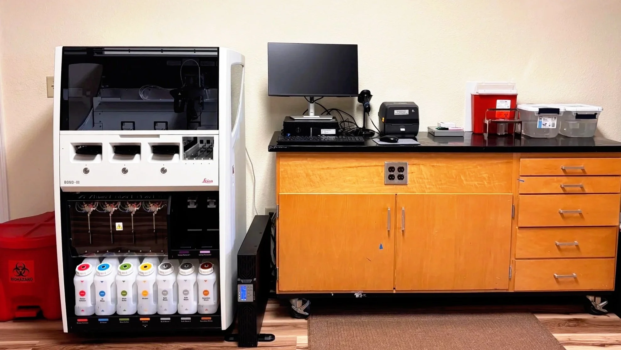Medical laboratory setup with a Leica Bond III on the left, a computer and label printer on a laboratory benchtop and organized supplies on the right.