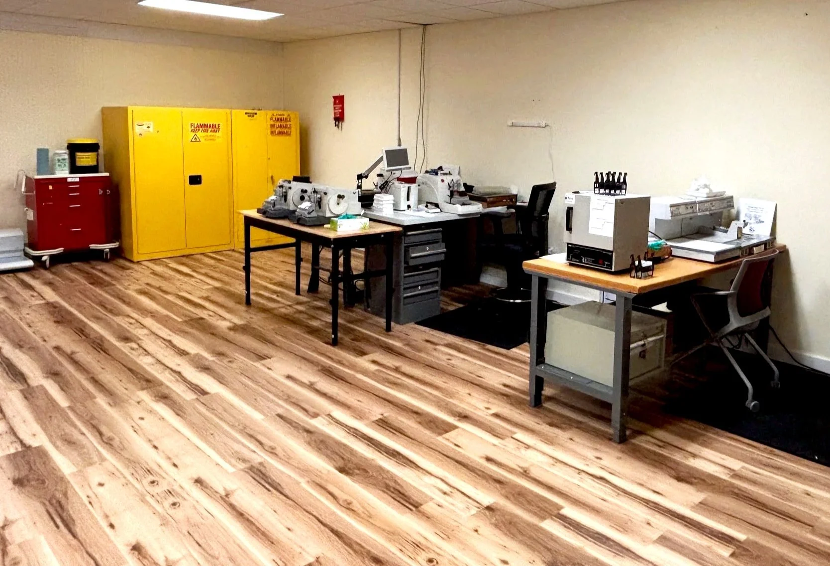Laboratory with faux wooden flooring, yellow flammable cabinets,' worktables with microtomes, embedding center, slide printer, and chairs.