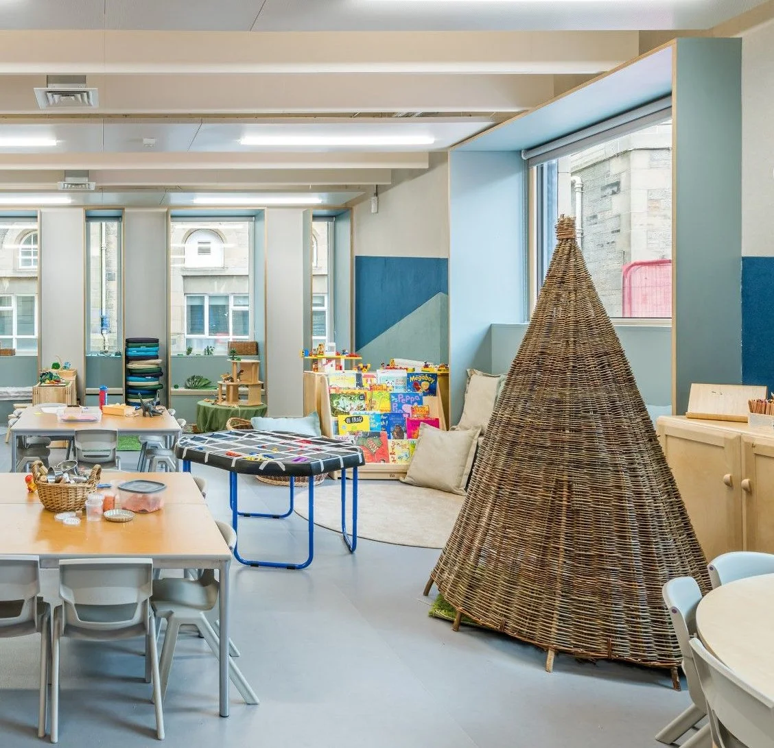 Bright classroom with tables, chairs, bookshelves, and a woven teepee tent, featuring large windows and colorful toys and books.