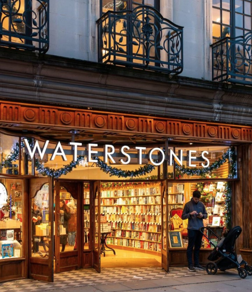 Exterior of Waterstones bookstore decorated with Christmas garlands, with open doors revealing shelves of books inside and a person with a stroller outside using a smartphone.