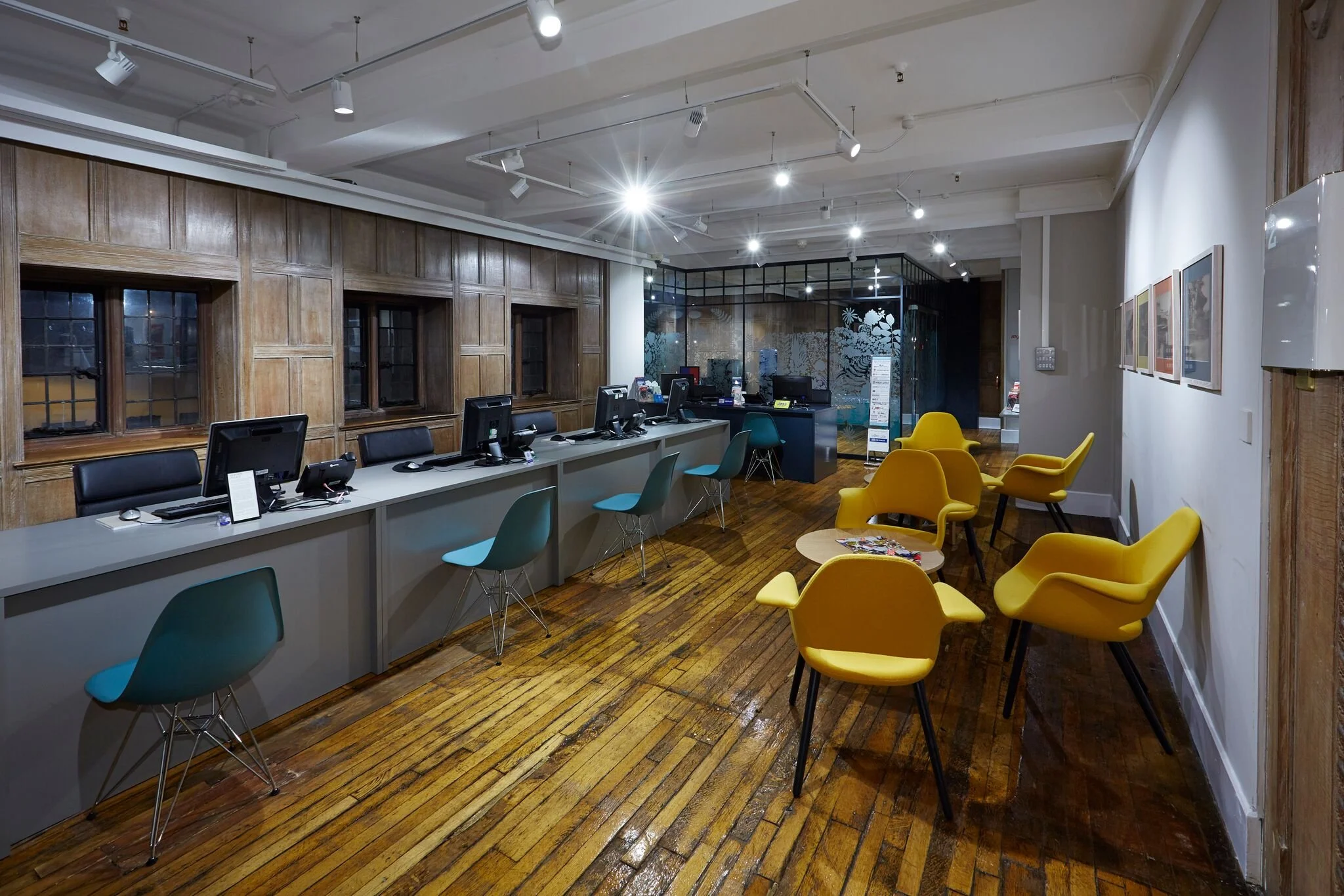 Interior of a modern reception area with a long desk, six computer workstations, blue and yellow chairs, wooden floor, and a decorative glass partition in the background.
