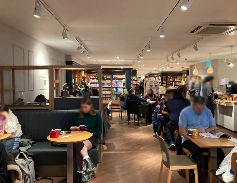 People sitting at tables and reading in a cozy cafe or bookstore with bookshelves in the background.